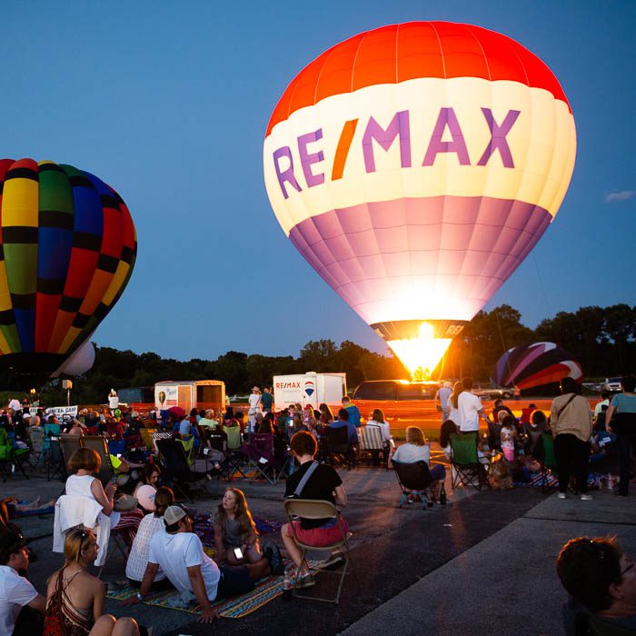 A crowd of people sitting on the ground watching hot air balloons.